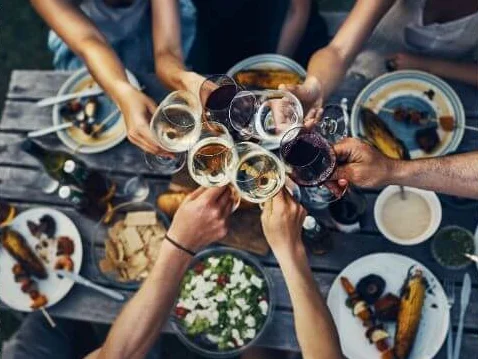 Manos que brindan con copas de vino blanco y tinto sobre una mesa de madera con platillos