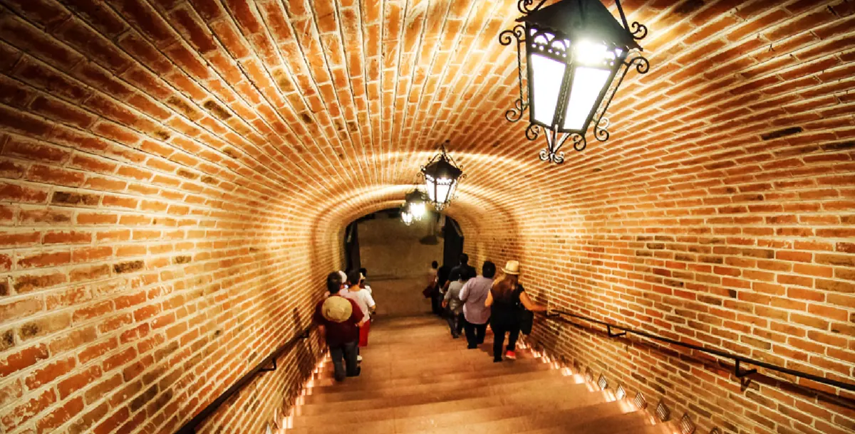 Visitantes bajan por la escalera abovedada de ladrillo hacia la cava de Finca Sala Vivé, en Ezequiel Montes, alumbrada con faroles de hierro.