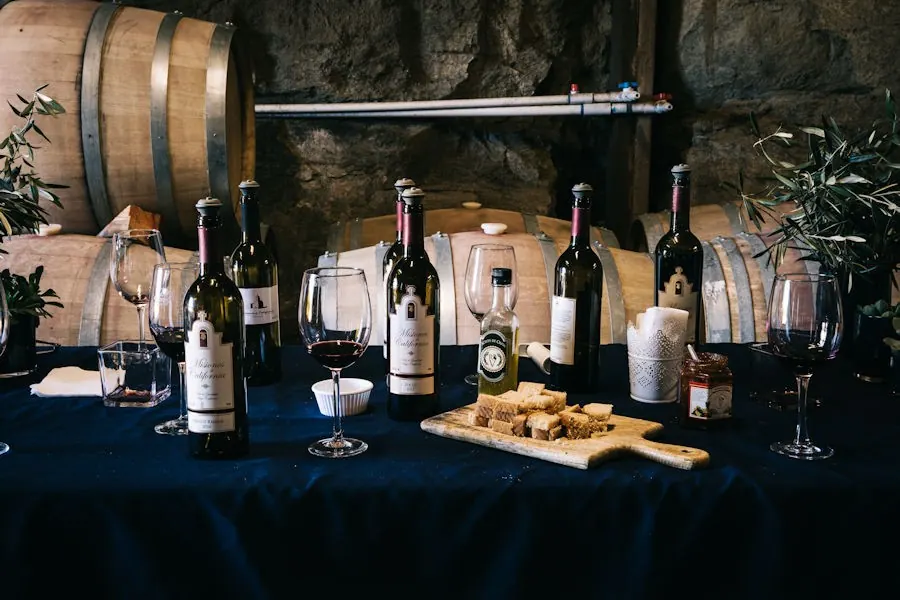 Mesa con mantel azul oscuro en la cava de Misiones de California: botellas de la casa, copas con vino tinto, cubos de pan tostado sobre una tabla de madera, aceite de oliva y un frasco de mermelada, con barricas al fondo.