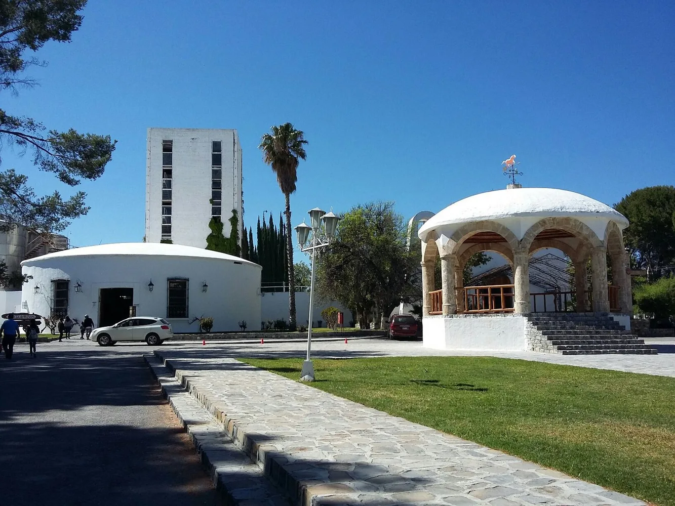 Kiosco de cantera con cúpula blanca y veleta; al fondo, un edificio circular encalado y una torre blanca entre palmeras y cipreses, en la hacienda de Casa Madero, en Parras, Coahuila.