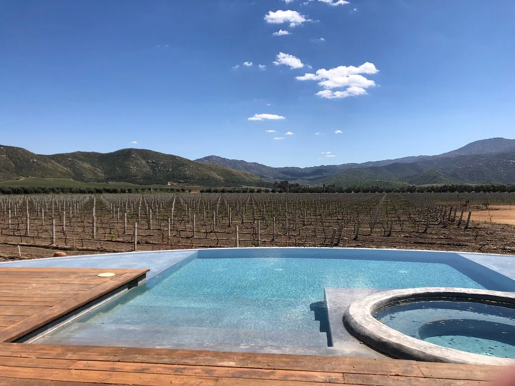 Alberca y jacuzzi con deck de madera frente a un viñedo sin hojas y cerros, en Bruma, Valle de Guadalupe.